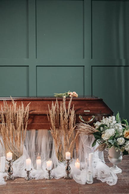 A floral tribute arranged on a wooden surface in front of a green paneled wall, featuring a collection of white roses, peonies, and lush greenery, complemented by tall, dried pampas grass for texture.