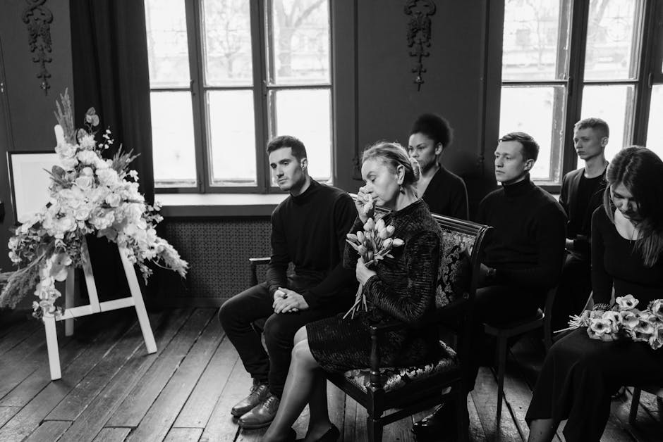 A group of six individuals seated indoors during a somber occasion, with a large funeral floral arrangement on a stand to the left. The arrangement features a variety of white flowers, including roses