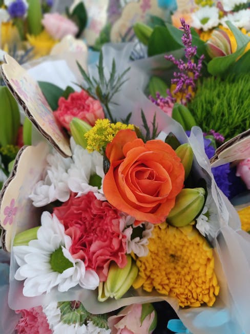 A vibrant floral bouquet featuring a prominent orange rose surrounded by white daisies, pink carnations, yellow chrysanthemums, and purple lavender sprigs. The flowers are arranged in a compact, round