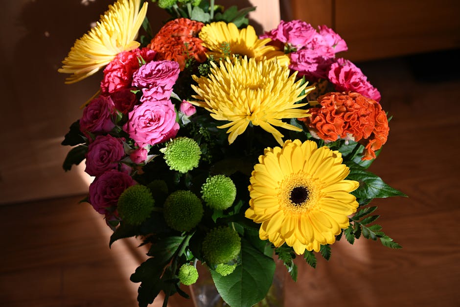 A vibrant floral arrangement featuring bright yellow gerbera daisies, pink roses, orange carnations, green button chrysanthemums, and yellow chrysanthemums, all arranged in a glass vase. The bouquet s