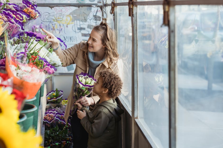 A woman and a young girl inside a flower shop, casually dressed, selecting a floral gift. The woman, smiling, is pointing toward a vibrant display of colorful flowers, including purple, red, and white