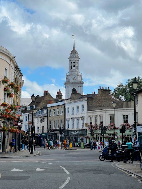The image depicts a bustling street scene in Wealdstone, with a prominent historic clock tower and church spire in the background under a partly cloudy sky. The street is lined with charming Victorian