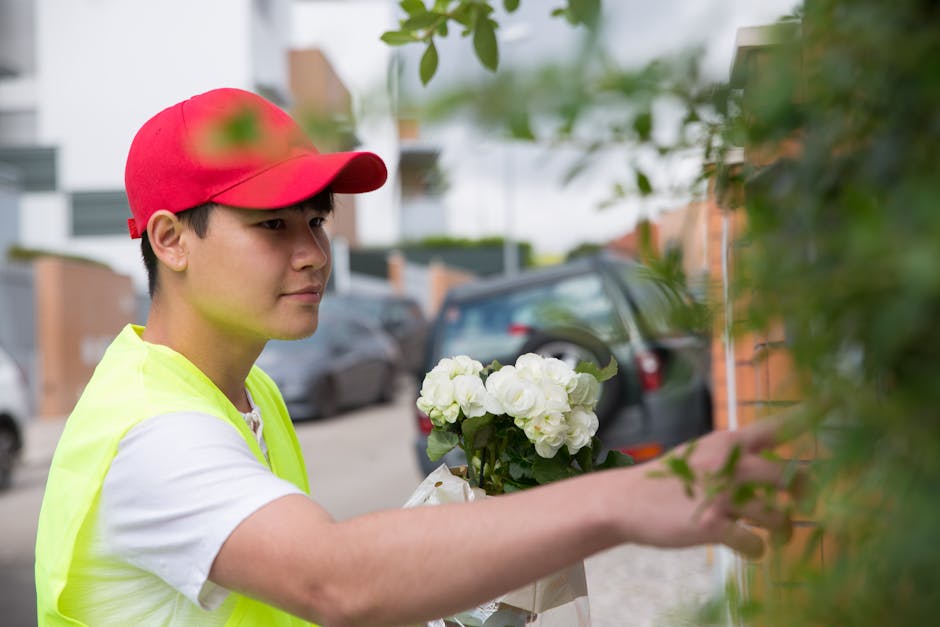 A young male florist in a red baseball cap and high-visibility yellow vest arranges a bouquet of fresh white roses and hydrangeas against a brick wall, with parked cars and modern residential building