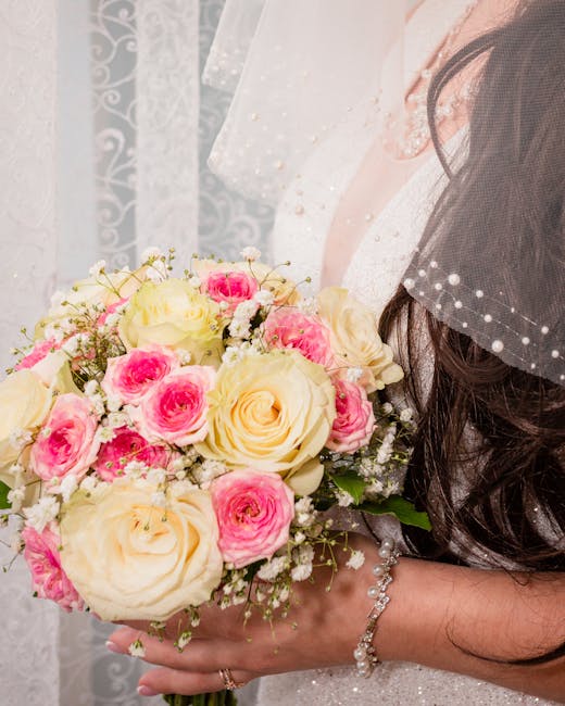 A bride holds a bouquet arrangement featuring large, creamy-white roses and vibrant pink roses, accented with delicate baby's breath and green foliage. The bouquet is gathered in a rounded, tightly cl