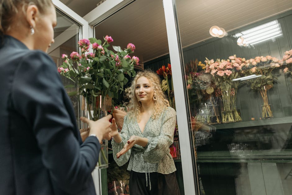 A woman with curly blonde hair wearing a light-colored floral blouse receives a bouquet of pink roses with green leaves from a floral shop assistant dressed in a dark jacket. The bouquet is wrapped in
