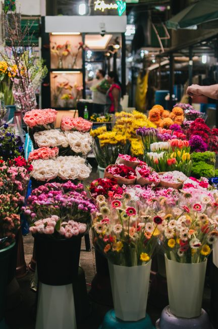 A vibrant floral display at a busy flower market, featuring a variety of fresh flowers arranged in tall white and black vases and buckets. The bouquet includes pink roses, white peonies, pink and whit