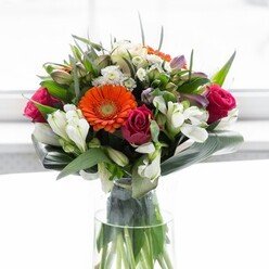 This exquisite floral arrangement stands as a testament to the artistry and heart found in Harrow Weald's beloved flower shops. An elegant gathering of creamy peach and fiery orange gerbera daisies draws the eye, each flower a symbol of cheerful exuberance, encircling deep brown centers that glow like lanterns against the restful green. Nestled nearby, bold magenta roses offer a sense of luxury and depth, their unfurling petals a nod to the enduring romance celebrated in our local community. White alstroemeria and occasional soft pink filler blossoms weave throughout, illuminating the bouquet with a fresh, garden-inspired touch-much like the tranquil green spaces around Harrow Weald. Carefully curved, broad green leaves wrap protectively around the base, echoing the nurturing nature of local florists and providing a grounding serenity amidst the bright blossoms. The arrangement sits proudly in a tall, clear glass vase, its vivid stems visible and adding to the bouquet's lively energy. Gentle daylight sweeps across, making each petal and leaf come alive in soft focus, mirroring the welcoming brightness of a window on a spring day. The bouquet is perfectly suited for joyful gatherings, special anniversaries, or moments of gratitude shared in Harrow Weald. Every element has been chosen with care, creating a composition that sings with optimism, beauty, and the spirit of thoughtful gifting.